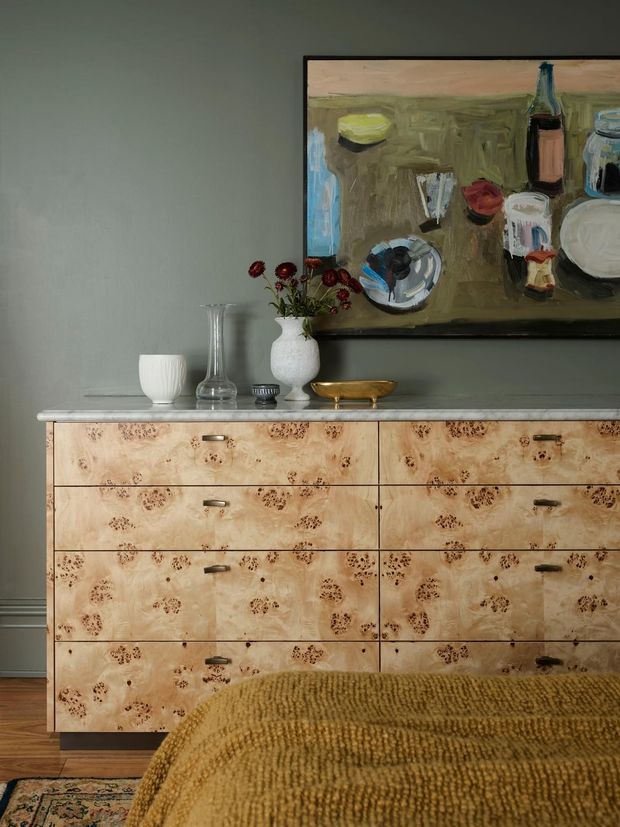 A moody bedroom vignette featuring a low timber chest of drawers with distinctive figured and patterned veneer, dark metal pulls, and a pale stone-like top. Decorative ceramics, glass vessels, a brass bowl, and a vase of flowers sit on the dresser beneath a large framed still-life painting. A textured mustard-brown throw covers the foreground bed, while the muted green-grey wall and dark timber flooring create a warm, refined atmosphere.