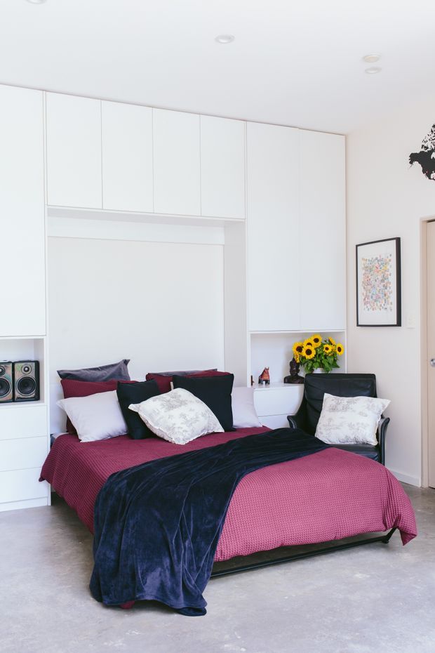 Bright contemporary bedroom with a double bed dressed in deep pink bedding and a navy throw, white built-in cabinetry spanning the walls, a black lounge chair, framed artwork, and small decorative items. The room has a light neutral palette, recessed ceiling lights, and a polished concrete floor.