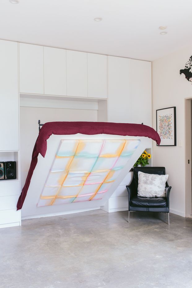 Bright contemporary bedroom with a partially raised wall bed, white built-in cabinetry, polished grey concrete flooring, and a black leather lounge chair. The bed features a colourful translucent panel underside and burgundy bedding, with framed artwork and a small floral arrangement adding accents.