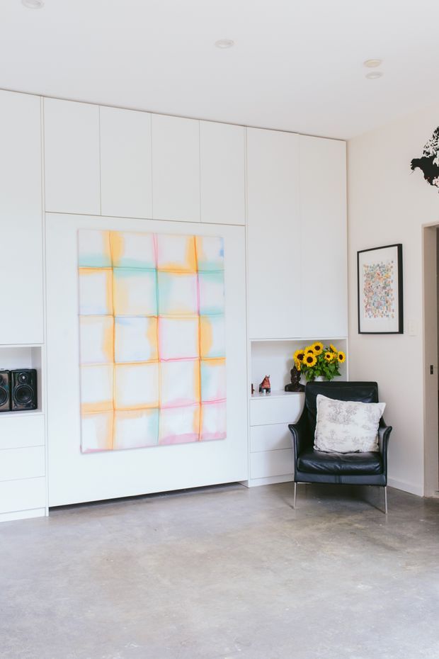 Bright minimalist living area with extensive white built-in cabinetry and open shelving, a large colourful abstract artwork, and a black leather lounge chair with a decorative cushion. The room has a smooth grey concrete floor, white walls and ceiling with recessed downlights, framed artwork, and a small arrangement of sunflowers. A doorway is visible at right.