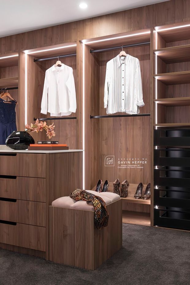 A refined walk-in wardrobe with extensive dark timber veneer cabinetry, open hanging bays, illuminated shelving, drawers, shoe storage, and a central upholstered island. Clothing and accessories are displayed throughout, with integrated warm LED strip lighting and a plush dark carpet floor.