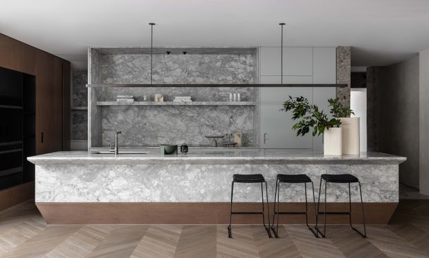 A refined contemporary kitchen featuring a large veined marble island with integrated sink and three black bar stools. The kitchen includes a matching marble splashback and open shelving, dark timber cabinetry, pale grey cabinetry, black appliances, pendant lighting, and a warm herringbone timber floor.