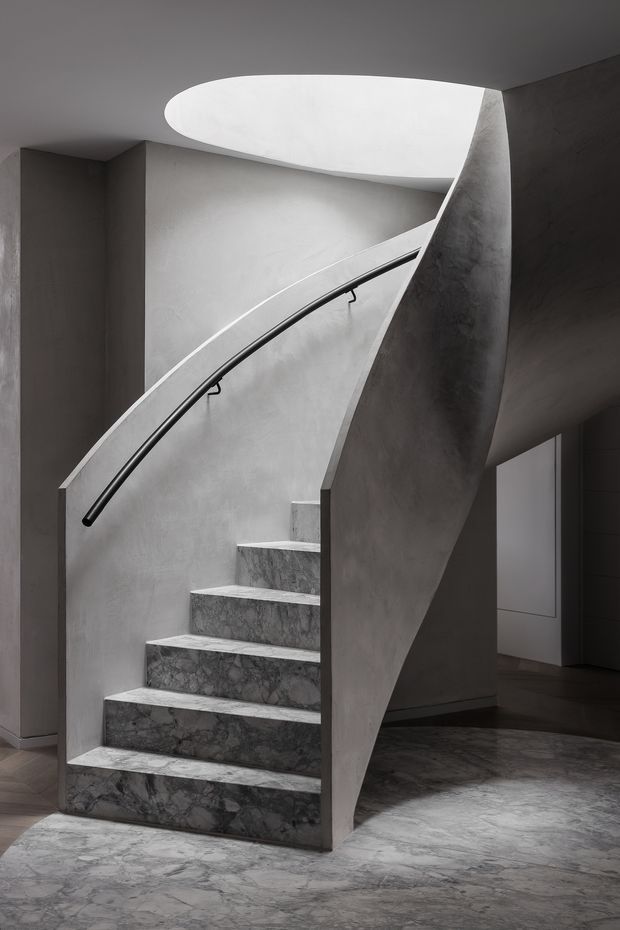 A sculptural curved staircase in a monochromatic interior, with grey marble-clad steps and flooring, smooth plastered walls, a dark curved handrail, and an elliptical skylight or ceiling opening bringing in soft natural light. The composition emphasises flowing geometry, shadow, and minimal detailing.