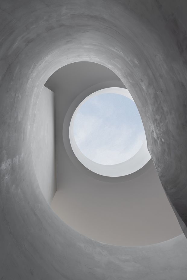 Upward-looking architectural detail of a sculptural curved stair or vaulted passage, with rough grey plaster walls forming an arched opening around a smooth white oval skylight. The composition is monochromatic and minimal, emphasizing organic geometry, diffuse daylight, and the contrast between textured and smooth surfaces.