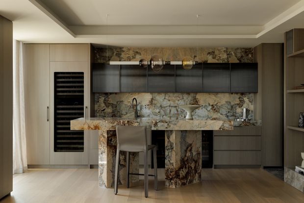 A refined contemporary kitchen with a compact marble-clad island and matching full-height marble splashback. Dark charcoal lower and upper cabinetry contrasts with pale timber-toned wall cabinetry and warm timber flooring. A built-in wine fridge, integrated sink and tap, open shelving, and suspended glassware are visible. The room has a layered recessed ceiling and soft, warm lighting.