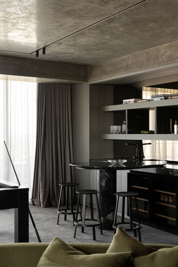 A dark, sophisticated open-plan living and bar area with a compact black stone island, integrated sink and matte-black tap, open shelving displaying glassware and books, and black cabinetry with wine storage. Several black stools surround the island. A low green sofa occupies the foreground, while full-height sheer curtains and heavy charcoal drapes frame a bright window. The ceiling and structural beam have a mottled concrete-like finish, creating a moody, refined contemporary interior.