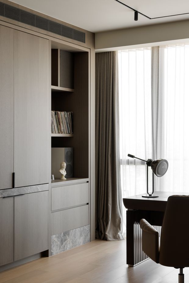 A refined compact home office or study corner with full-height light wood built-in cabinetry, open shelving, drawers, and a marble-look feature panel. A dark wood desk sits beside a large sheer-curtained window, paired with a sculptural upholstered office chair and black adjustable task lamp. The room has pale timber flooring, warm taupe walls, integrated linear air conditioning, and minimalist black track lighting.