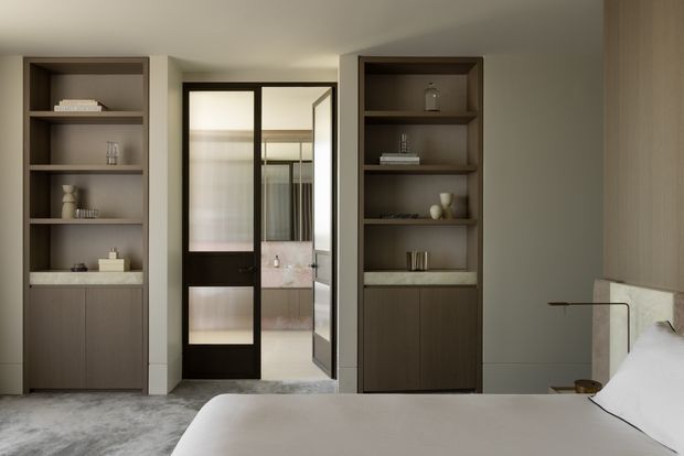 A refined contemporary bedroom viewed toward a central ensuite doorway, flanked by symmetrical dark built-in shelving and cabinetry. The room has a soft neutral palette, pale textured carpet, a low-profile bed with white bedding, warm taupe walls, and dark-framed glazed doors. A bathroom with a freestanding or built-in tub is visible beyond the doorway.