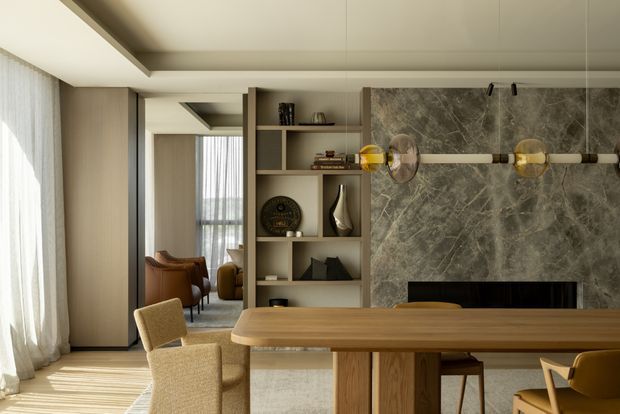 Refined contemporary dining and living space with a long timber dining table in the foreground, upholstered dining chairs, built-in timber shelving, and a dramatic grey stone fireplace feature wall. Floor-to-ceiling sheer curtains provide soft daylight, while sculptural wall lighting and curated shelving add warmth and sophistication. A lounge area is visible beyond the open shelving.