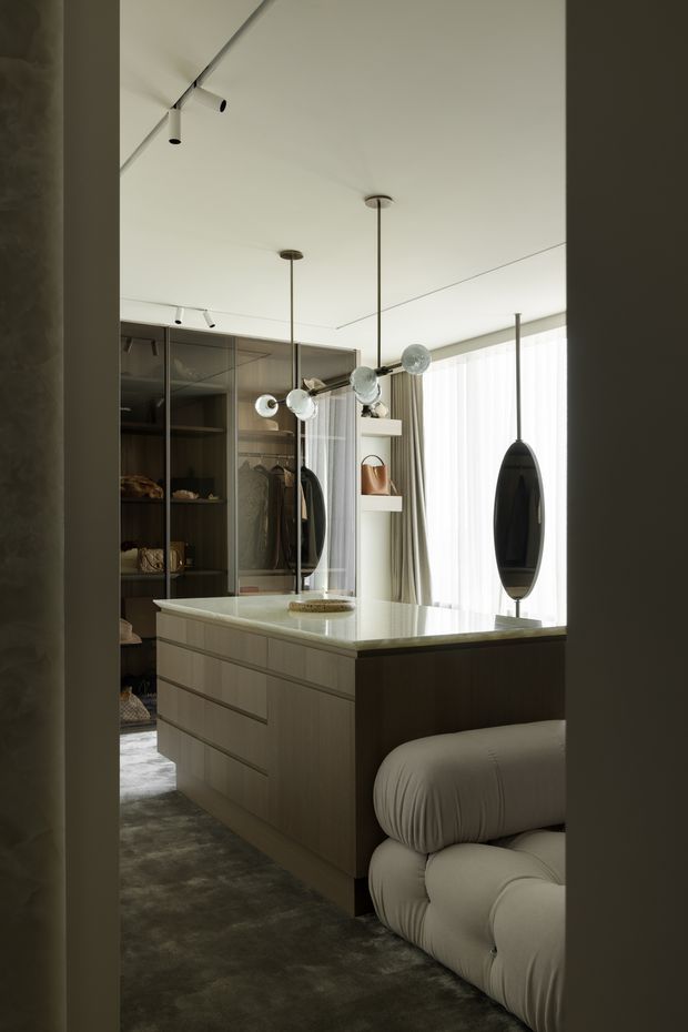 A refined walk-in dressing room or wardrobe area centred on a large island of dark timber drawers with a pale stone benchtop. Floor-to-ceiling glass-fronted wardrobes line the rear wall, displaying clothing and accessories, while sculptural pendant lights hang above the island. A plush cream upholstered seat occupies the foreground beside full-height sheer curtains and glazing. The palette is muted and warm, with soft layered lighting and luxurious contemporary detailing.