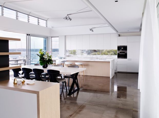 Bright, open-plan contemporary kitchen and dining area with floor-to-ceiling glazing overlooking a coastal or waterfront setting. The room features white full-height cabinetry, a pale timber island and dining furniture, polished concrete-look flooring, black dining chairs, and a long horizontal splashback window. High-level louvred windows and minimalist track lighting enhance the airy interior.