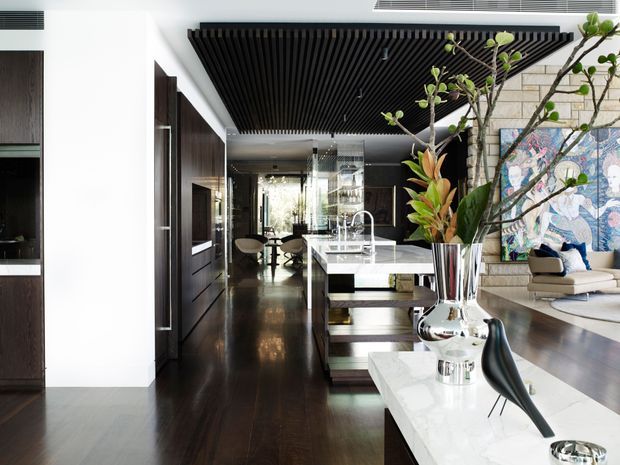 A sophisticated open-plan contemporary interior featuring a kitchen island with a pale stone benchtop, dark timber cabinetry and flooring, and a dramatic black slatted ceiling feature. The space flows into a dining and living area with sculptural furniture, large botanical arrangements, a stone-clad feature wall, and colourful artwork. Glossy reflective surfaces and extensive glazing create a refined, high-contrast atmosphere.