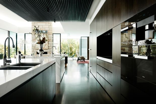 A spacious contemporary kitchen with a large white stone island, integrated sinks and modern taps, dark timber cabinetry, and a full-height bank of built-in appliances. A dramatic black slatted ceiling contrasts with the bright glazing, pale stone feature wall, and polished dark floor. The open-plan space extends toward a light-filled living area with garden views.