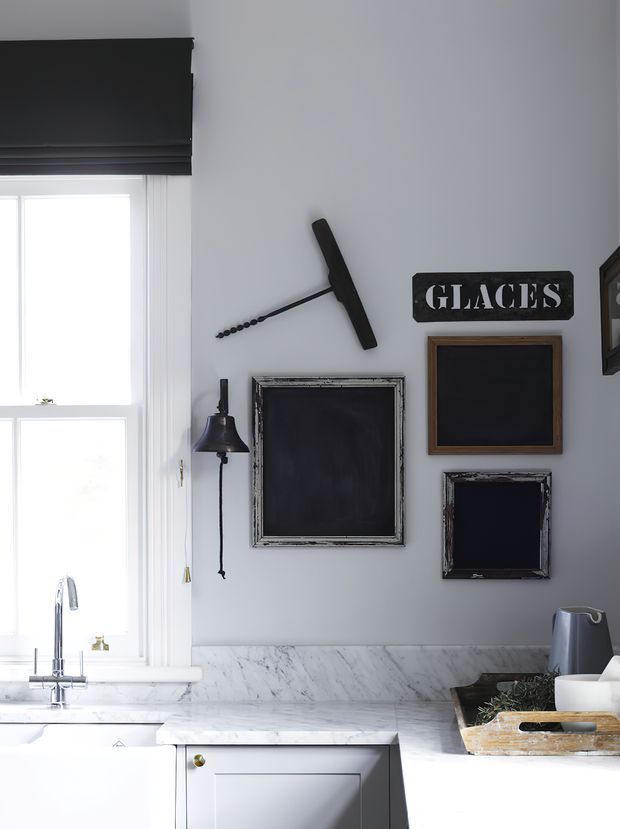 Bright, compact kitchen corner with a white marble countertop and backsplash, white cabinetry, a stainless steel gooseneck faucet and sink beneath a window, and dark framed wall decor including a sign reading “GLACES.” A black roller blind and dark decorative accents provide contrast against pale grey walls.
