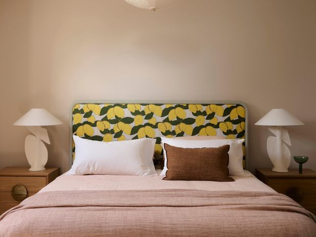 A warmly styled bedroom with a large upholstered bedhead featuring a bold botanical pattern in olive green, mustard yellow and cream. The bed is dressed in white pillows and layered blush-pink and brown bedding, flanked by timber bedside tables and sculptural white table lamps. The walls are warm beige, and a subtle ceiling light is visible above.