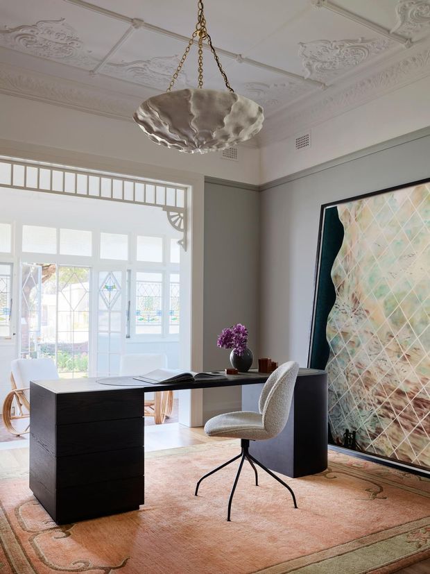 Elegant home office in a heritage-style interior, featuring a black minimalist desk, upholstered swivel chair, oversized framed artwork, ornate plaster ceiling details, decorative ceiling rose, and a sculptural pendant light. Glazed doors and windows with leadlight-style details open toward a bright adjoining space. A large patterned rug adds warmth and softness.