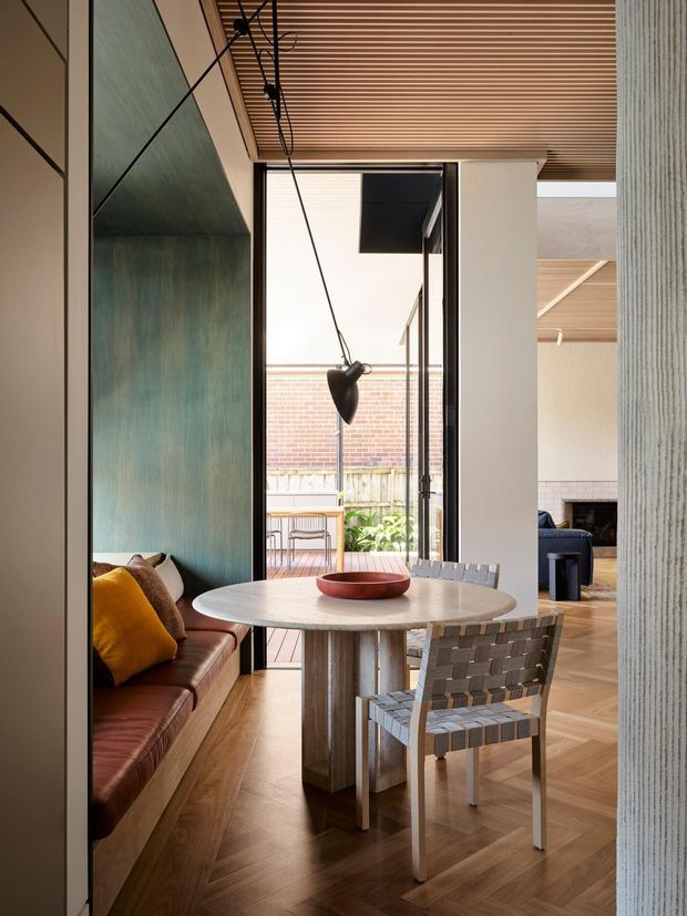 A refined dining nook with a round stone-topped table, built-in leather bench seating, and a woven dining chair. The space features warm herringbone timber flooring, a richly textured teal wall panel, vertical textured wall finish, and a slatted timber ceiling. Full-height glazing opens toward a small courtyard, while a suspended black pendant light hangs above the table. A living area with fireplace is visible beyond.