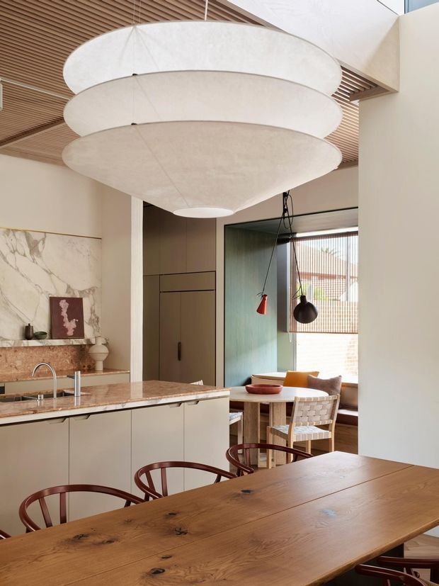 A warm, refined open-plan kitchen and dining area with a large timber dining table in the foreground, a pale stone kitchen island with integrated sink, full-height cabinetry, marble-look wall slab, and a dining nook beyond. The space features sculptural layered pendant lighting, a slatted timber ceiling, timber-framed furnishings, and muted green and neutral finishes.