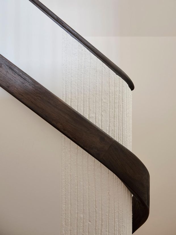 Close-up architectural detail of a curved stairway with dark stained timber handrails and balustrade elements wrapping around a textured white plaster or concrete central wall. The composition is minimal and sculptural, with soft neutral lighting and no visible furnishings.