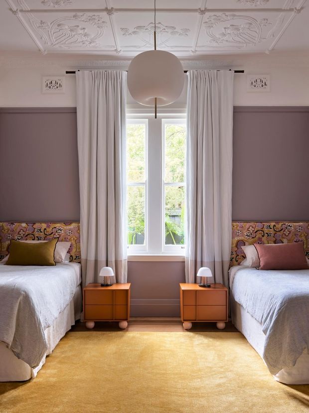 Symmetrical twin bedroom with two single beds, colourful upholstered headboards, matching orange bedside cabinets and lamps, a central tall window with sheer curtains, and a large warm yellow rug. The room features mauve lower walls, white upper walls, an ornate decorative ceiling, and a sculptural spherical pendant light.