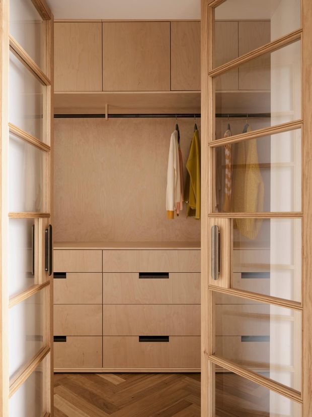 A compact walk-in wardrobe viewed through open timber-framed glazed doors. The space features light timber veneer cabinetry with upper cupboards, open hanging rails, drawers with dark recessed pulls, integrated handles, and a herringbone timber floor. Clothing is visible on the hanging rails, and the overall design is warm, refined, and highly organized.