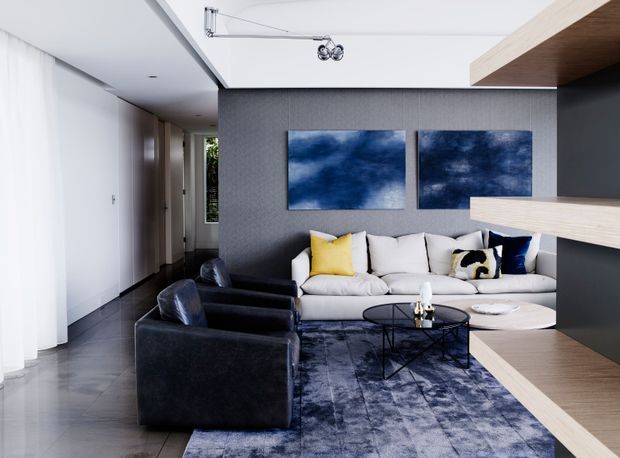 Contemporary living room with a light sectional sofa, dark leather armchairs, a round glass coffee table, and a large blue-grey patterned rug. The room features dark textured wall panelling, white walls and ceiling, built-in light timber shelving or cabinetry, recessed downlights, and blue abstract artwork.