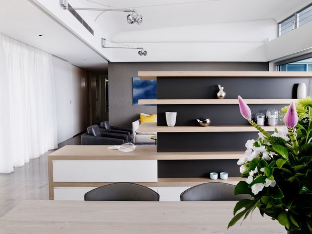 Bright contemporary open-plan living and dining area viewed from behind a pale timber dining table. A substantial built-in timber-and-dark shelving partition separates the dining zone from a lounge with dark armchairs and a pale sofa. White sheer curtains, a white ceiling with modern track-style lighting, light flooring, and large glazing create an airy, refined interior. A large floral arrangement occupies the foreground.