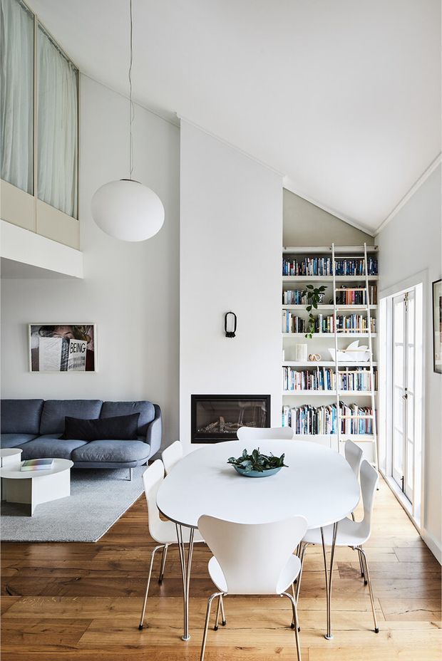 Bright, high-ceiling open-plan living and dining area with a large white oval dining table and sculptural white chairs in the foreground. A grey sofa, pale rug, fireplace, extensive built-in book shelving, pendant light, timber flooring, and tall glazed doors create a clean contemporary interior with abundant natural light.