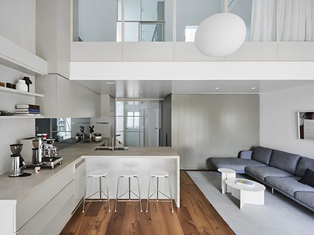 A bright, open-plan contemporary interior combining a white kitchen with a peninsula island and three bar stools, a compact living area with a grey sectional sofa and white nesting coffee tables, and warm timber flooring. White cabinetry, integrated shelving, a large spherical pendant light, and a partial mezzanine or upper-level void create a clean, airy feel.