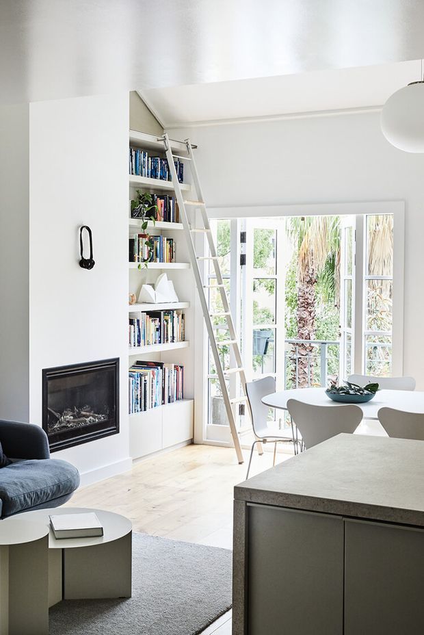 Bright, refined open-plan interior combining a living area, dining space and kitchen. White built-in floor-to-ceiling bookshelves with a library ladder frame a fireplace wall, while large glazed doors open to greenery outside. A pale kitchen island occupies the foreground, with light timber flooring, neutral furnishings and a sculptural pendant light.