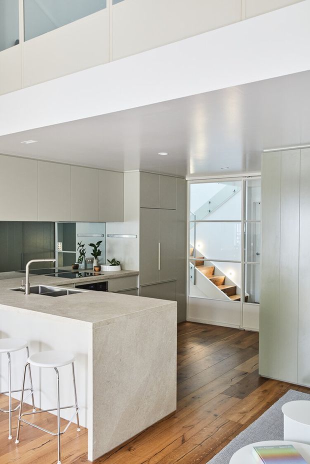 Bright, contemporary kitchen with extensive pale grey built-in cabinetry, a large stone island with integrated sink, white bar stools, timber flooring, and a glazed internal door opening toward a stairwell. A partial upper-level mezzanine or void is visible above the kitchen, contributing to an airy, open layout.
