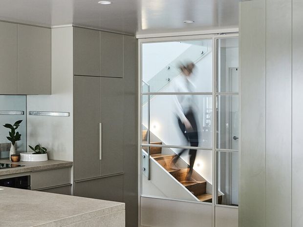 A bright, compact contemporary kitchen with integrated grey cabinetry, a pale stone island benchtop, stainless-steel refrigerator and white walls. A glazed internal partition reveals a timber staircase and a blurred person beyond, while recessed ceiling lights provide clean illumination.