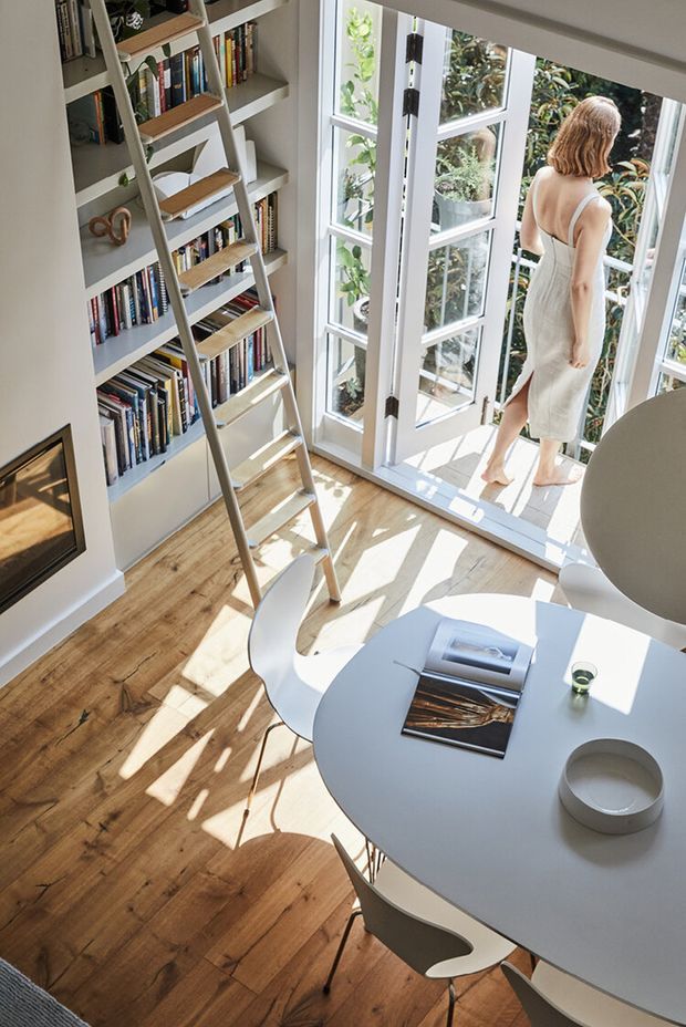 Bright, airy dining and reading corner with a built-in white bookshelf filled with books, an integrated rolling ladder, and a round white dining table with contemporary chairs. Full-height white-framed glazed doors open onto lush greenery, allowing strong sunlight to cast shadows across the warm timber floor. A partial wall-mounted screen or artwork is visible at left.