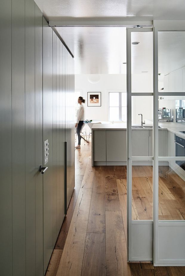 A bright contemporary kitchen viewed from a hallway or entry area, with full-height grey-green cabinetry along the left wall, a central island, white benchtops, timber flooring, and a glazed metal-framed partition door. The kitchen opens toward a light-filled dining or living area with a person in the background.