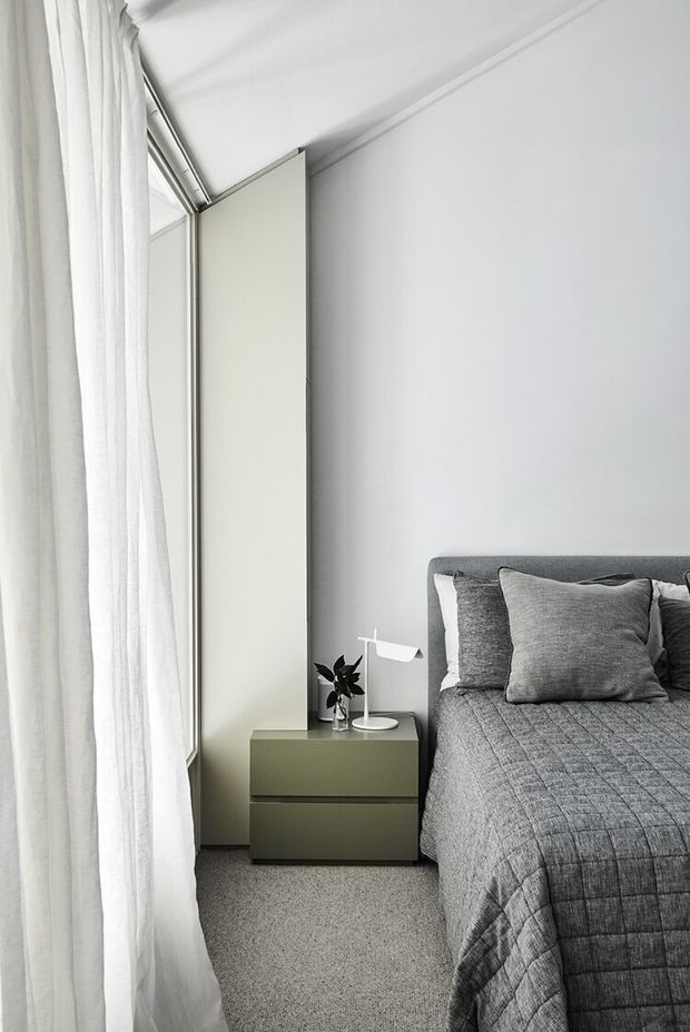 A compact, minimalist bedroom with a grey upholstered bed, layered grey bedding and pillows, a muted olive-green bedside cabinet, white modern wall lamp, pale walls, and full-height sheer curtains beside a tall window. The room is softly lit and predominantly neutral, with a calm, refined atmosphere.