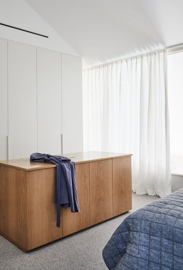 A bright, minimalist bedroom with a low, warm timber-finished island dresser or storage unit in the foreground, white built-in wardrobes along the wall, sheer full-height curtains over a large window, and a partially visible bed with a blue quilt. The room has a clean white palette, soft natural light, and understated contemporary detailing.