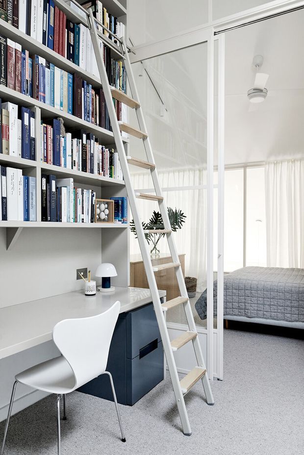 Bright, compact home office integrated beneath a full-height built-in bookshelf, with a white desk, sculptural white chair, dark blue drawer unit, decorative lamp, and rolling library ladder. A glass-partitioned bedroom with a grey quilted bed is visible beyond, with sheer curtains and abundant natural light.