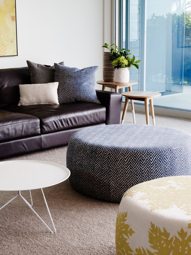 A bright contemporary living room featuring a dark leather sectional sofa, patterned cushions, a large blue-grey chevron upholstered ottoman, white nesting coffee tables, and a floral upholstered pouf. A potted plant sits on a small timber side table beside full-height glazed windows, with neutral carpet and white walls.