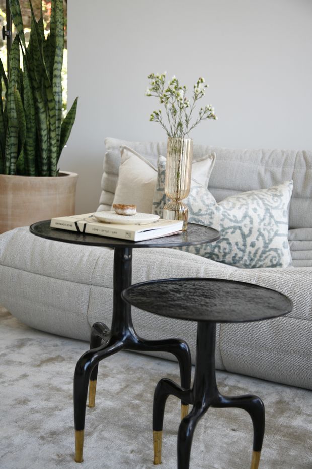A softly styled living room vignette featuring a pale textured sofa with patterned neutral cushions, two sculptural black side tables with brass-toned feet, a clear decorative vase, books, and a tall potted snake plant. The palette is neutral and refined, with a light rug and minimal grey wall backdrop.