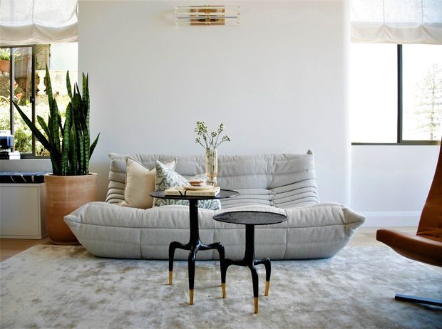 Bright, refined living room with a large cream upholstered sectional sofa, sculptural black-and-brass nesting tables, a pale area rug, tall indoor plants, and expansive windows with soft Roman shades. A simple wall-mounted brass light adds a subtle decorative feature, while a sculptural brown lounge chair is partially visible at right.