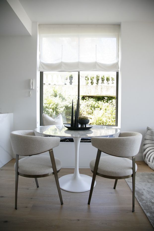 Bright, refined interior corner featuring a small round white pedestal table positioned by a large window, with two upholstered curved dining chairs. A white Roman shade filters daylight, while pale timber flooring, white walls, neutral furnishings, and a grey rug create a calm, minimalist setting. A sculptural black centerpiece sits on the table, and a sofa is partially visible at the right edge.