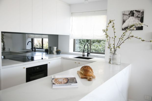 Bright contemporary white kitchen with a large island, integrated white cabinetry, a dark stone or composite benchtop, black tapware and appliances, and a mirrored splashback reflecting the workspace. A window provides garden views, while a vase of branches and framed artwork add restrained decorative touches.