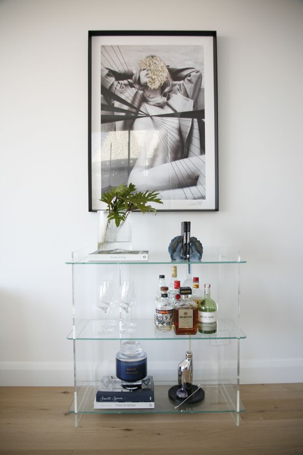 A styled interior vignette featuring a transparent glass drinks trolley or bar cabinet against a pale wall. The shelving holds wine and spirit bottles, cocktail glasses, books, and decorative objects, with a small leafy plant in a glass vase on top. A large black-framed monochrome artwork hangs above, and the unit stands on a light timber floor.