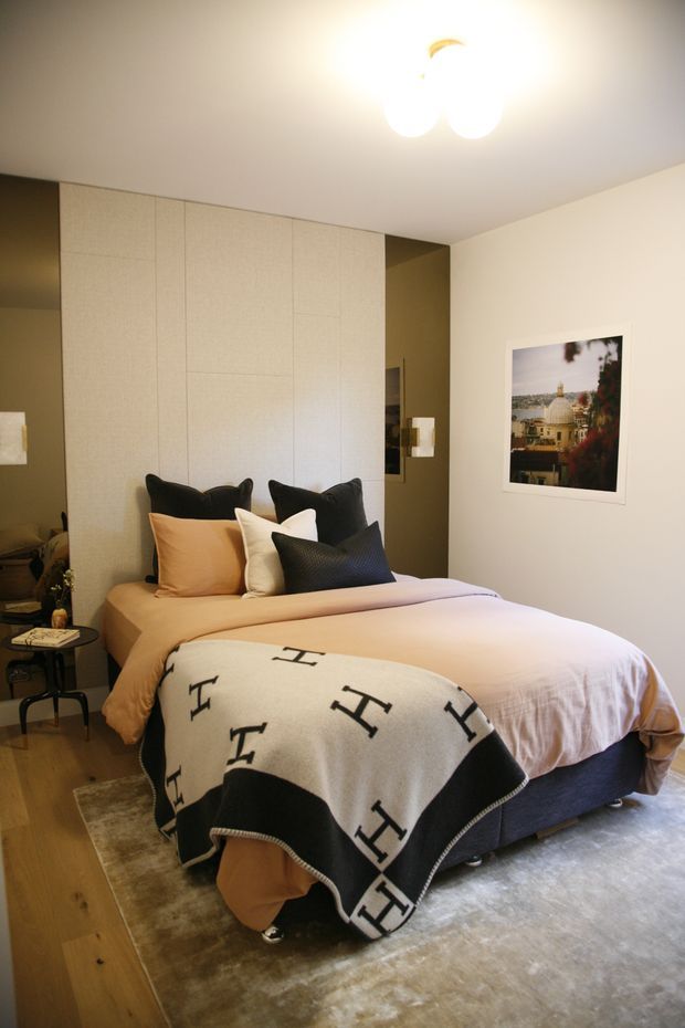 A warmly lit contemporary bedroom featuring a large upholstered bed with pale bedding, layered black, cream and ochre cushions, and a folded black-and-white patterned throw. Full-height textured light wall panelling forms a feature backdrop behind the bed, with dark mirrored or glossy joinery at the sides. Timber flooring, a large neutral rug, a small bedside table, and framed artwork complete the refined, cozy interior.