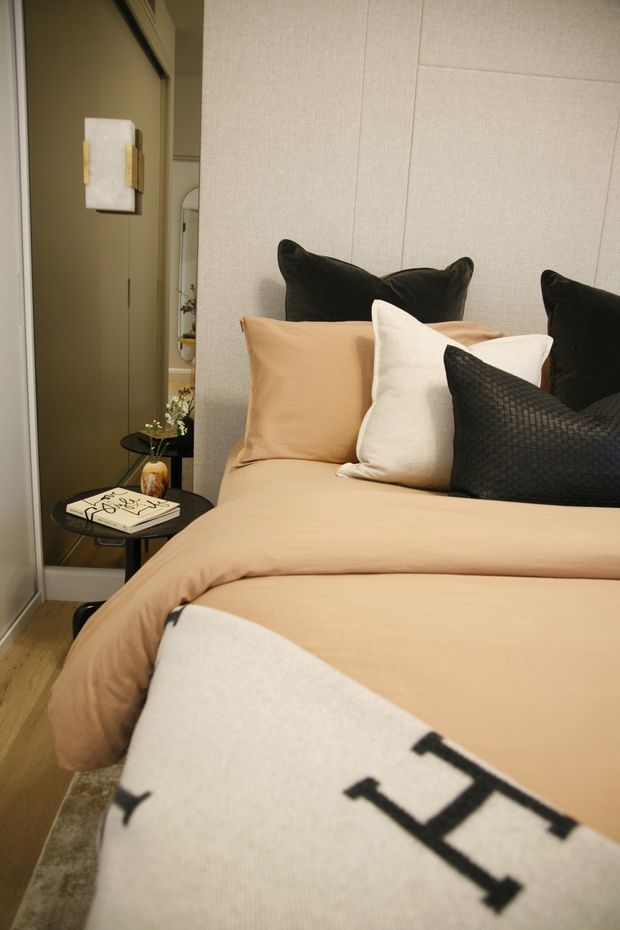 A warmly styled bedroom viewed close to the bed, featuring a tan upholstered or paneled wall, beige bedding, black and cream decorative pillows, a small black bedside table, timber flooring, and a narrow mirrored or reflective panel beside the bed. The room has a refined, cozy contemporary look with layered neutral textiles and dark accents.