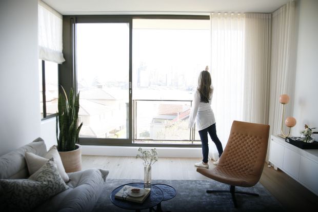 Bright contemporary living room with floor-to-ceiling windows, sheer curtains, a grey sofa, round glass coffee table, sculptural leather lounge chair, low white built-in cabinetry, and a tall indoor plant. The room overlooks a dense urban or suburban setting and has a calm, airy atmosphere.