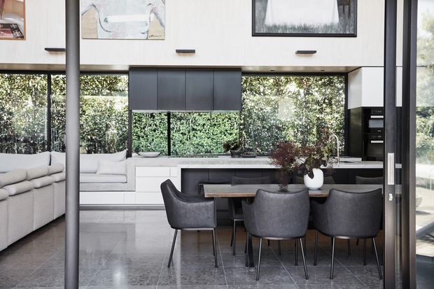 A bright, contemporary open-plan kitchen and dining area with a large island, dark upholstered dining chairs, pale cabinetry, extensive windows, and polished grey flooring. A living room with a light sectional sofa is visible to the left. The space features high ceilings, black-framed glazing, and integrated appliances.