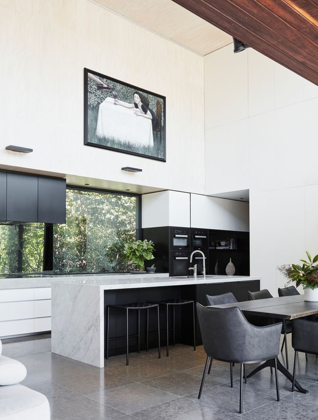 A sophisticated open-plan kitchen and dining area with tall white cabinetry, matte black integrated appliances, a veined stone island with black bar stools, and a dark dining table with upholstered chairs. Large windows overlook greenery, while a timber-lined ceiling feature and framed artwork add warmth and character.
