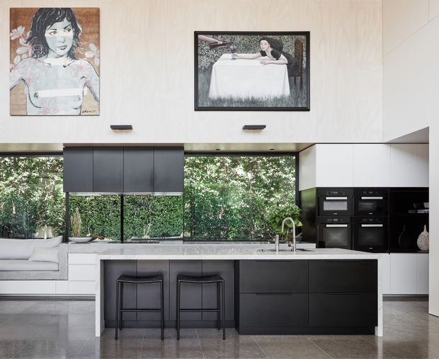 Contemporary open-plan kitchen with a large black island, integrated appliances, white cabinetry, dark stools, and expansive glazing overlooking greenery. Light timber wall panelling, polished grey flooring, and framed artwork create a refined, gallery-like interior.