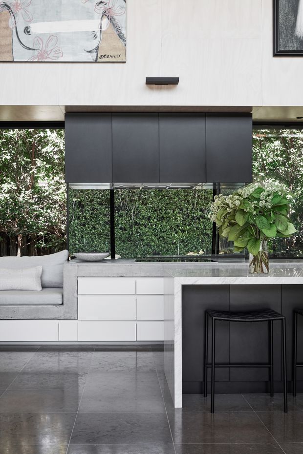Contemporary open-plan kitchen and living area with a monochrome palette. The kitchen features a large marble-look island with dark integrated cabinetry and black stools, white lower cabinetry, dark overhead cabinets, and extensive glazing overlooking dense greenery. A built-in upholstered window seat and artwork add softer residential elements. Large grey stone-look floor tiles complete the space.